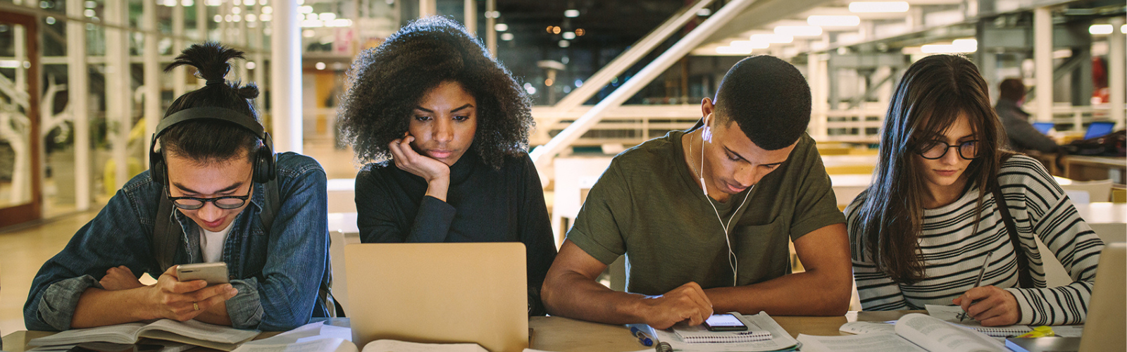 Four students studying using books, computers, pen and paper, and mobile devices.